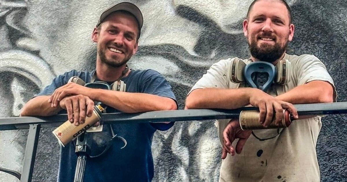 Two men in work gear pose in front of a graffiti-covered wall, both smiling. One holds a spray can.