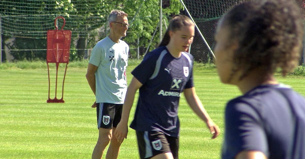 Ein Mann mit Brille und weißem Shirt steht neben einem Mädchen in einem blau-weißen Fußballtrikot. Sie befinden sich auf einem Feld mit einem Netz im Hintergrund.