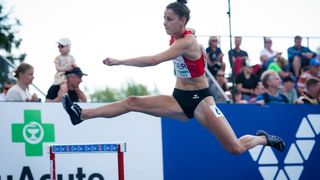 Eine Athletin in roter und schwarzer Sportkleidung springt über eine Hürde. Zuschauer schauen im Hintergrund zu.