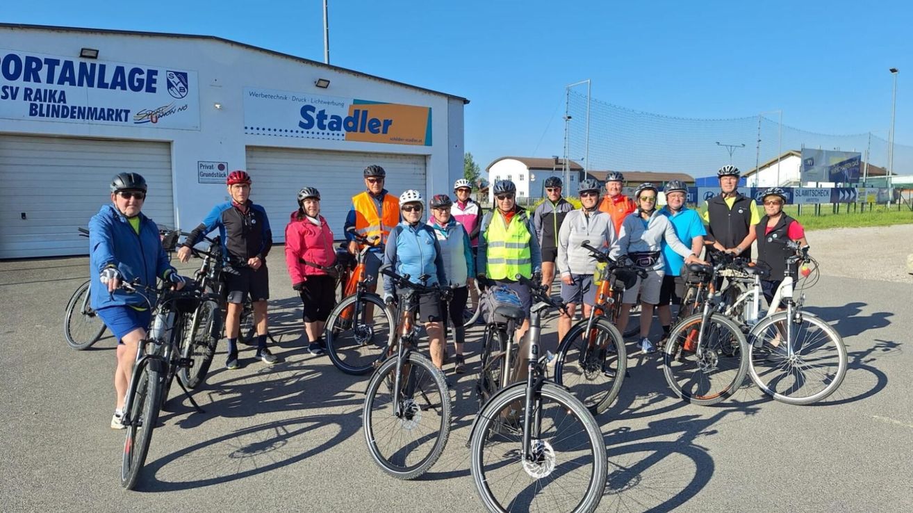 Eine Gruppe von Radfahrern mit Helmen und verschiedenen Ausrüstungsgegenständen posiert für ein Foto vor einem Gebäude mit einem Schild, auf dem 'Stadler' steht.