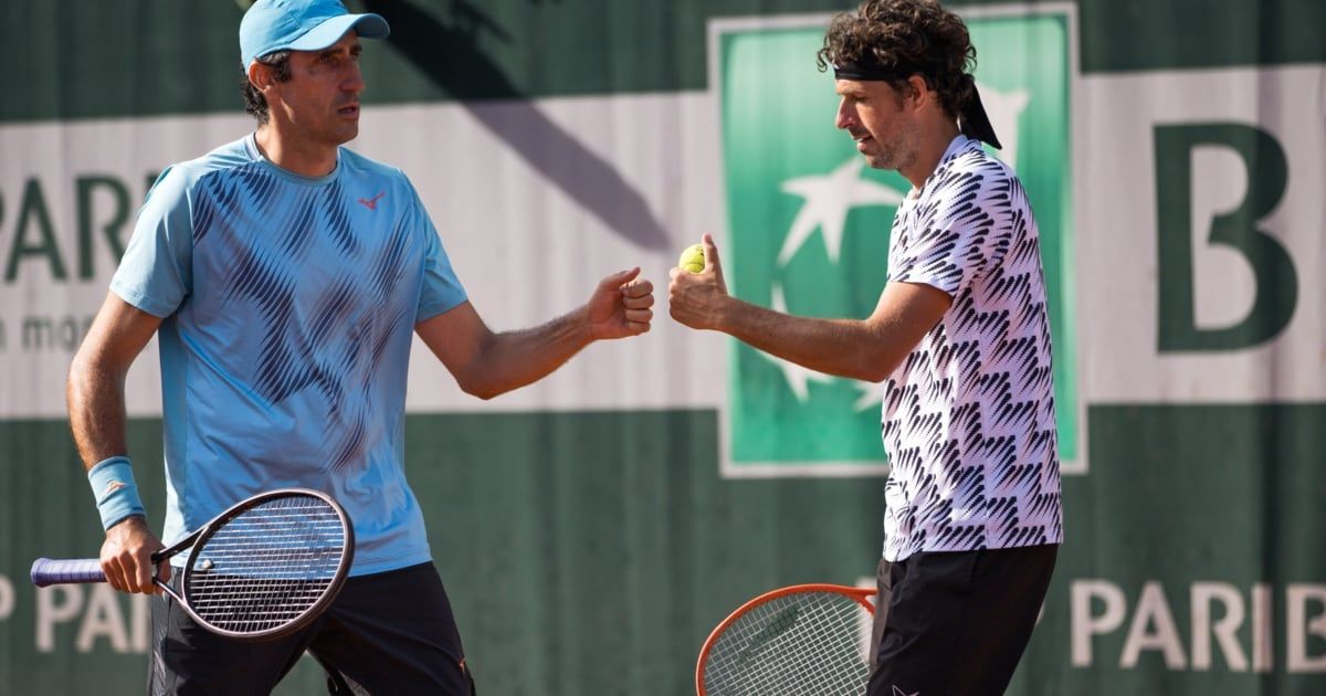 Zwei männliche Tennisspieler in Sportbekleidung stehen auf einem Tennisplatz, einer hält einen Tennisball, beide sehen einander an.