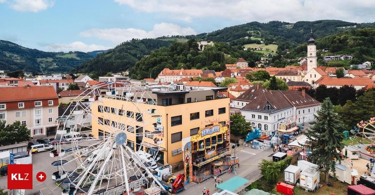 Luftaufnahme einer Stadt mit farbenfrohen Gebäuden, Bergen und einem großen Riesenrad. Menschen gehen herum und genießen das Jahrmarktgelände.