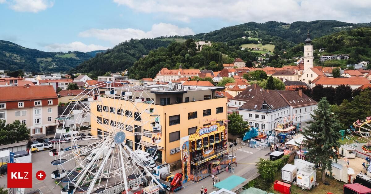 Luftaufnahme einer Stadt mit farbenfrohen Gebäuden, Bergen und einem großen Riesenrad. Menschen gehen herum und genießen das Jahrmarktgelände.