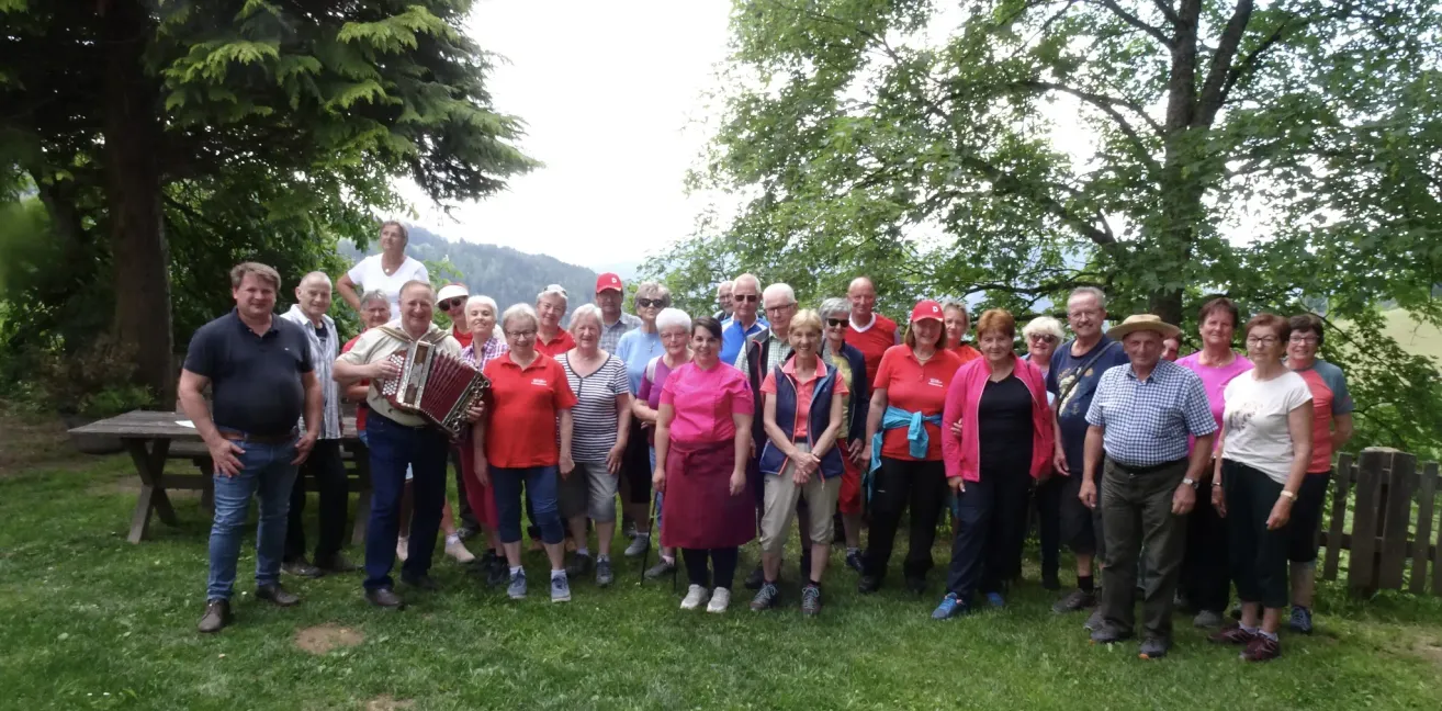 Eine Gruppe von Menschen in roten Shirts steht in einem Park und lächelt für ein Foto. Ein Mann spielt Akkordeon, während andere Sonnenbrillen und Hüte tragen. Sie sind von Bäumen umgeben.