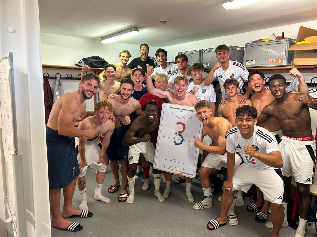 Eine Gruppe junger Fußballspieler posiert für ein Foto in einer Umkleidekabine, einige tragen Uniformen, andere Freizeitkleidung. Ein Spieler hält ein Whiteboard mit einer Fußballstrategie.