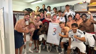 Eine Gruppe junger Fußballspieler posiert für ein Foto in einer Umkleidekabine, einige tragen Uniformen, andere Freizeitkleidung. Ein Spieler hält ein Whiteboard mit einer Fußballstrategie.