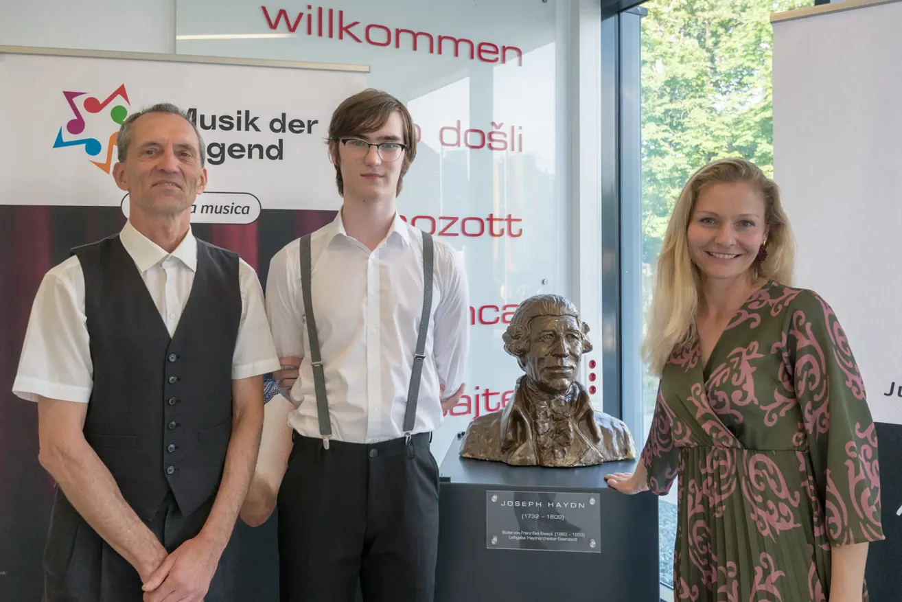 Drei Personen stehen neben einer Büste von Joseph Haydn und tragen formelle Kleidung. Der Mann auf der linken Seite trägt eine schwarze Weste. Der Mann in der Mitte trägt eine Trägerhose. Die Frau auf der rechten Seite trägt ein grünes Kleid.