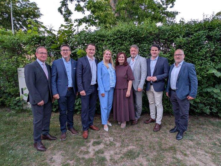 Eine Gruppe von sieben Fachleuten in Geschäftskleidung, die vor einem grünen Busch stehen und für ein Foto lächeln.