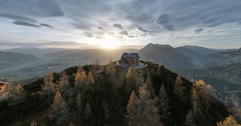 Ein Haus auf einem Berggipfel, umgeben von Herbstbäumen und einem bewölkten Himmel. Die Sonne geht hinter den Bergen in der Ferne unter.