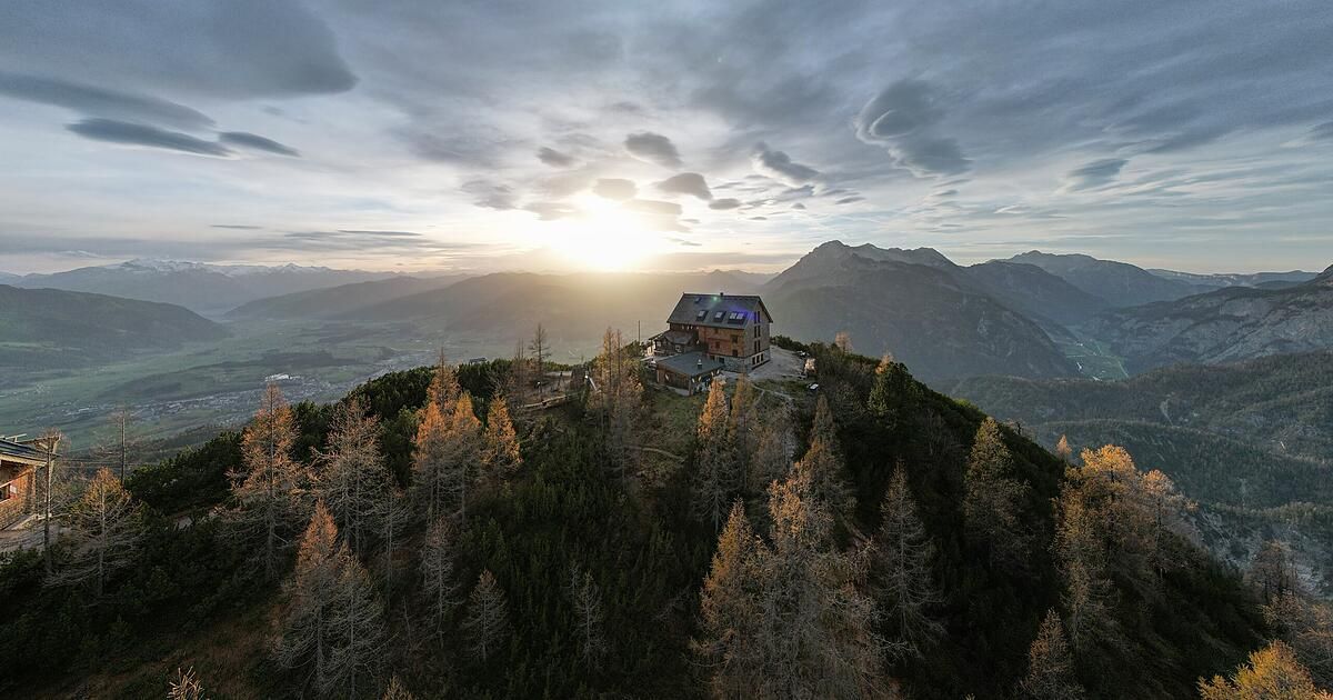 Ein Haus auf einem Berggipfel, umgeben von Herbstbäumen und einem bewölkten Himmel. Die Sonne geht hinter den Bergen in der Ferne unter.