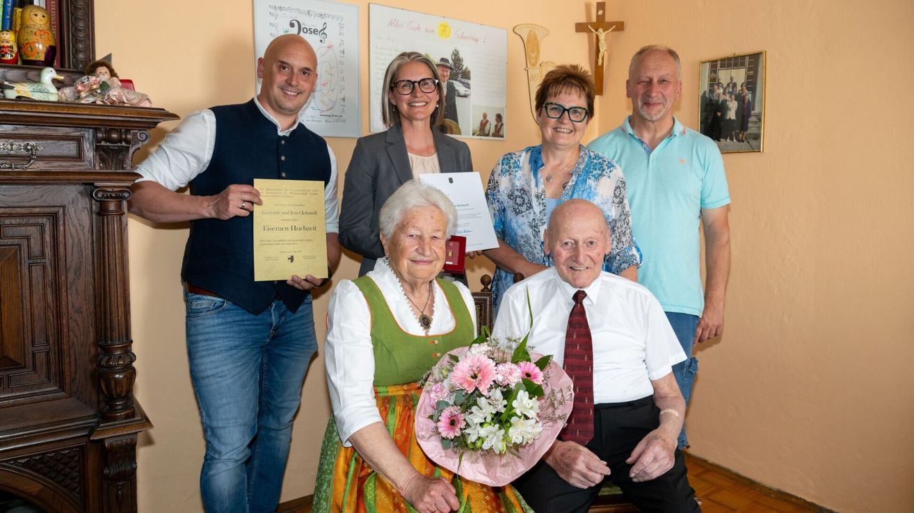Ein älteres Paar mit einer Auszeichnung und Blumen sitzt umgeben von Familie. Hinter ihnen befinden sich Poster mit einem Musiknotensymbol, einem Kreuz und Bildern.