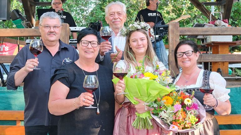 Eine Gruppe von Menschen bei einer Outdoor-Veranstaltung. Eine Frau mit Blumen in den Haaren hält einen Blumenstrauß und ein Weinglas. Ein Mann hinter ihr hält ein Weinglas. Andere Menschen und ein Musiker sind im Hintergrund.