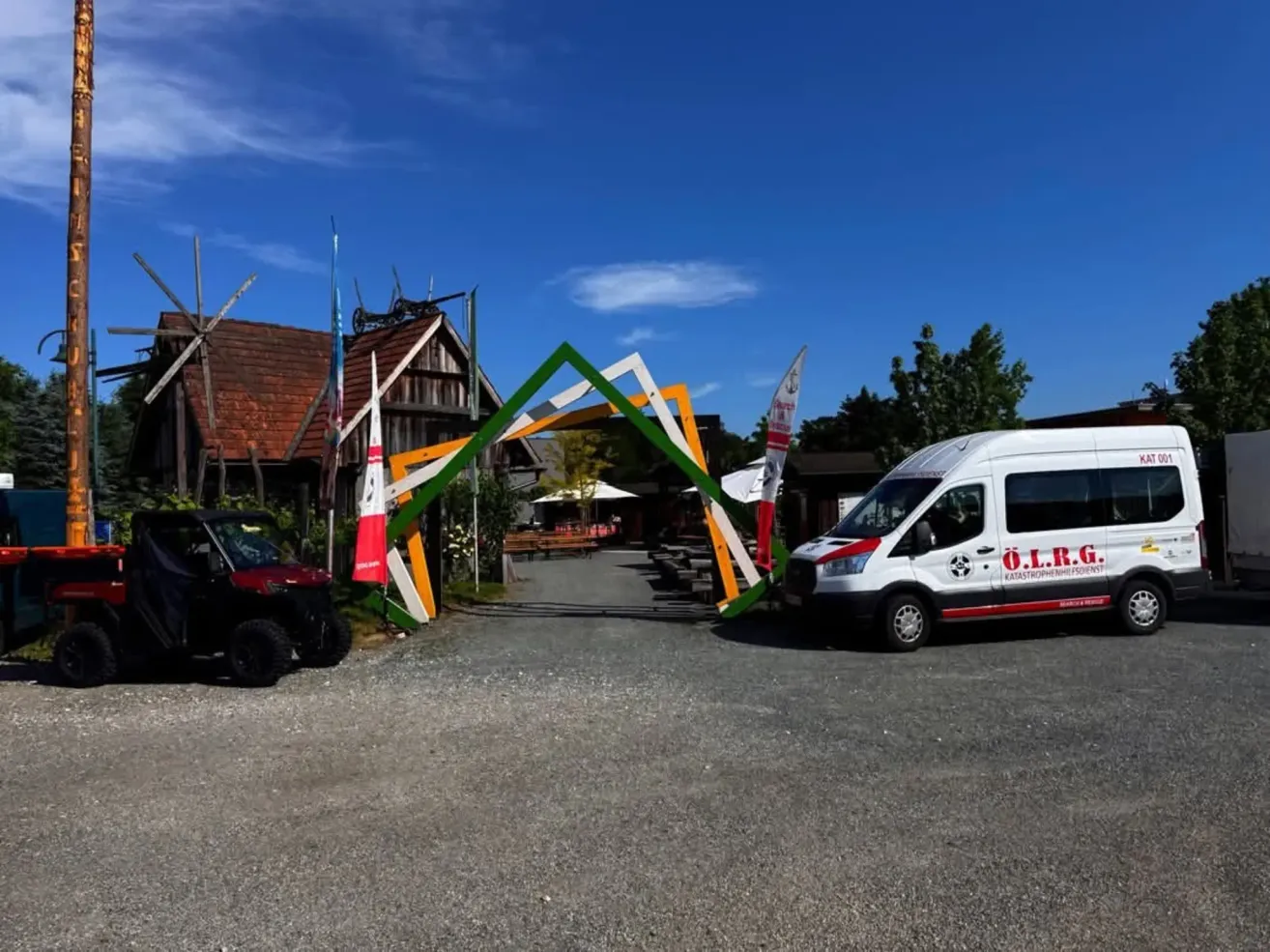 Ein farbenfroher Bogeneingang in ein ländliches Gebiet mit einem geparkten Van und einem Fahrzeug, einem Haus mit Flaggen und einem bewölkten Himmel.