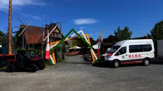 Ein farbenfroher Bogeneingang in ein ländliches Gebiet mit einem geparkten Van und einem Fahrzeug, einem Haus mit Flaggen und einem bewölkten Himmel.
