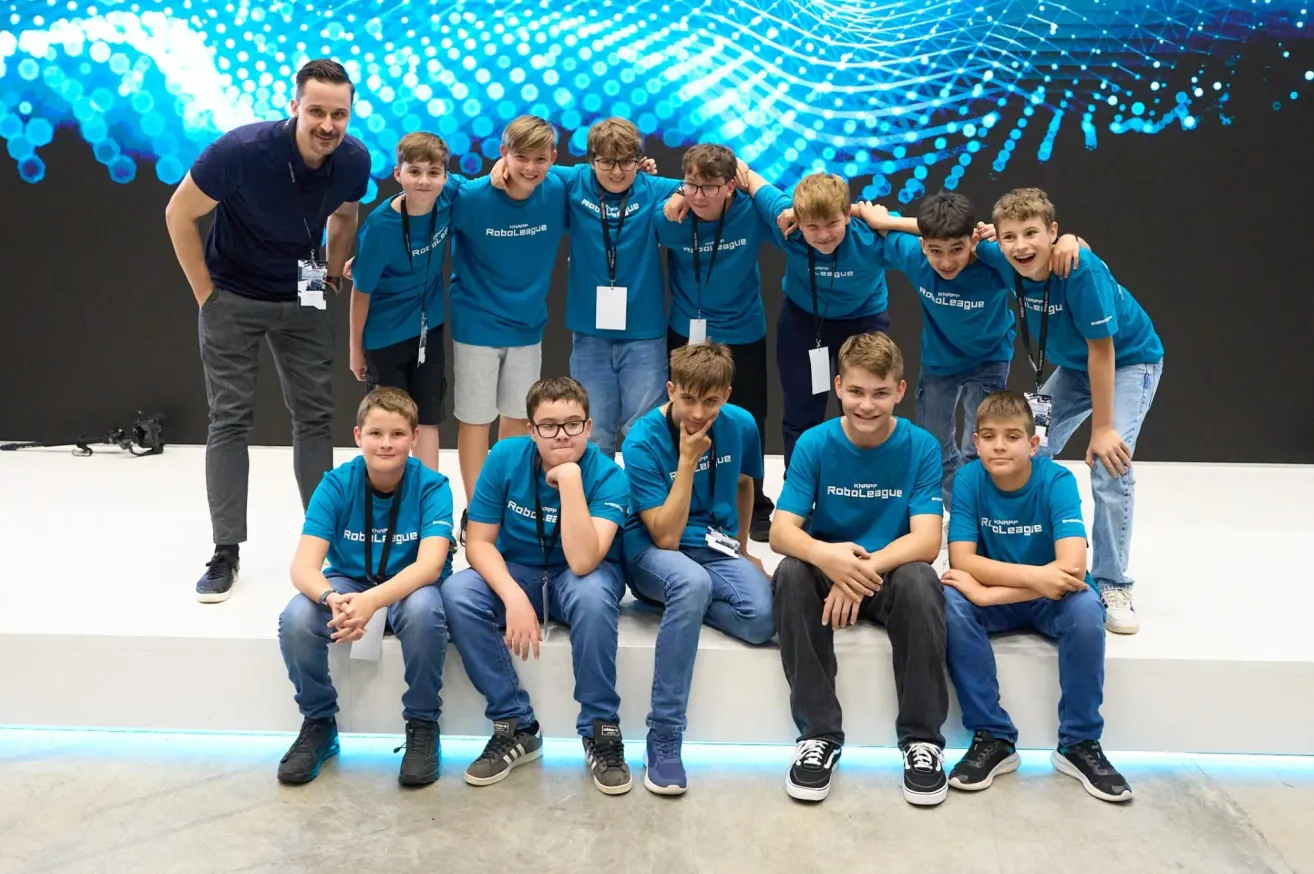 Eine Gruppe Jungen in blauen Shirts mit 'RoboLeague' darauf, die auf einer weißen Bank sitzen. Ein Mann steht hinter ihnen mit einem lächelnden Gesicht.
