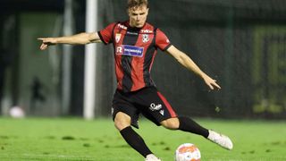 Ein Fußballspieler in rot-schwarzem Trikot mit 'RHOMBERG' auf der Brust tritt den Ball auf einem Rasenfeld.