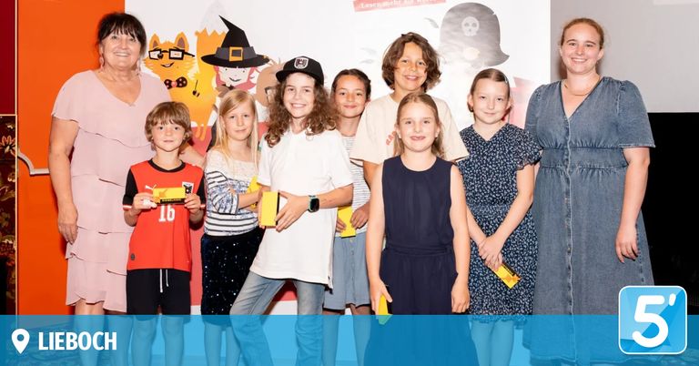 Several children and adults stand together, smiling and posing for a photo with yellow booklets in their hands in front of a white background with drawings.
