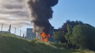Ein Gebäude in Brand sendet dicken schwarzen Rauch aus, umgeben von Bäumen und einem Zaun, vor einem blauen Himmel.
