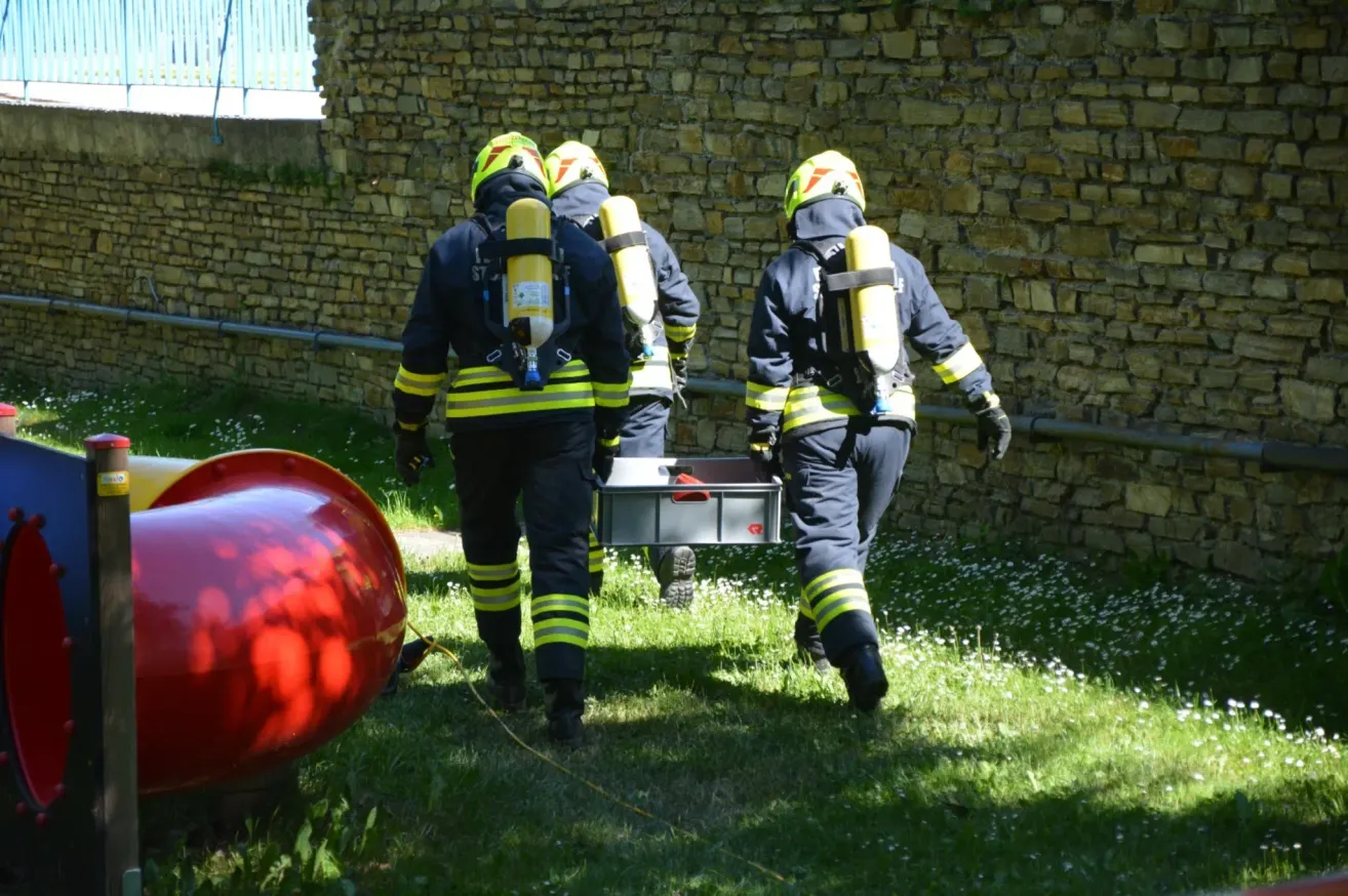 Drei Feuerwehrleute in Schutzausrüstung, die Gerät und einen Behälter tragen, gehen auf einer Rasenfläche in der Nähe einer Steinmauer.