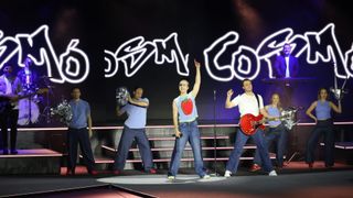 Eine Band tritt auf der Bühne mit einer Scheinwerferbeleuchtung auf. Ein Mann winkt mit der Hand und ein anderer spielt eine rote Gitarre. Zwei weitere sind dahinter.