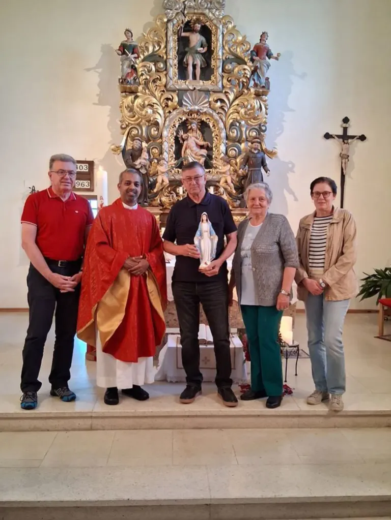 Fünf Menschen stehen in einer Kirche und posieren für ein Foto. Ein Priester hält eine Statue, und ein Kreuz ist an der Wand.