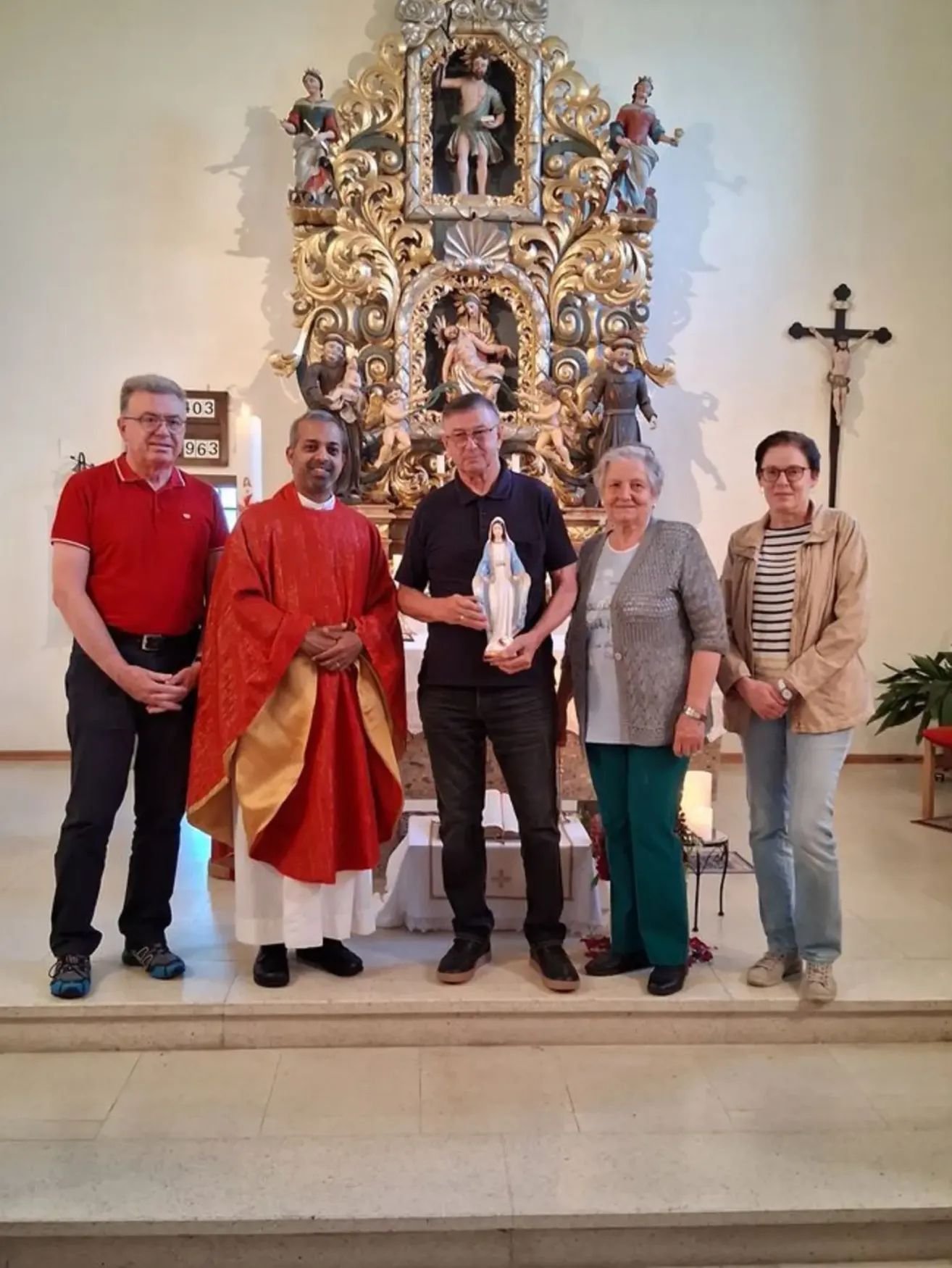 Fünf Menschen stehen in einer Kirche und posieren für ein Foto. Ein Priester hält eine Statue, und ein Kreuz ist an der Wand.