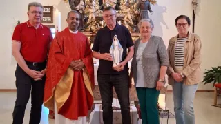 Fünf Menschen stehen in einer Kirche und posieren für ein Foto. Ein Priester hält eine Statue, und ein Kreuz ist an der Wand.