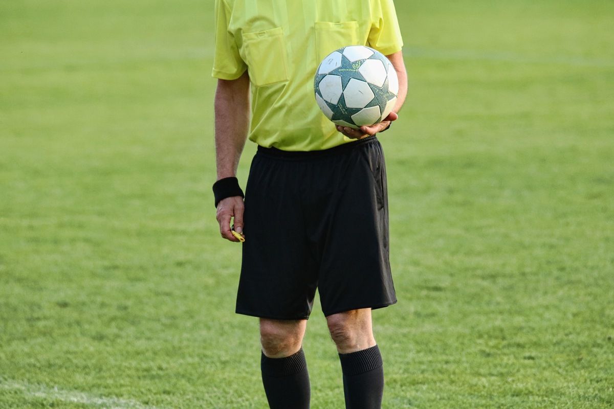 Ein Fußballschiedsrichter hält einen Fußball in der Hand, gekleidet in einem neongelben Shirt, schwarzen Shorts und schwarzen Socken. Er befindet sich auf einem grünen Spielfeld.