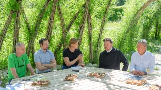 Vier Personen sitzen an einem Tisch im Freien, mit Bambuswänden hinter ihnen. Ein Mann hat gefaltete Arme, eine Frau gestikuliert während des Sprechens und ein Mönch hört zu. Weingläser und Essen stehen auf dem Tisch.