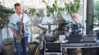 Zwei Männer spielen Instrumente, einer spielt Saxophon und der andere spielt am Keyboard. Sie stehen vor einer Glasscheibe mit Reflexionen eines Mannes und eines Tisches.