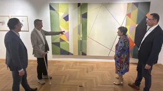 Ein Mann zeigt auf ein großes abstraktes Gemälde, während zwei Frauen und ein anderer Mann in einer Galerie in der Nähe stehen.