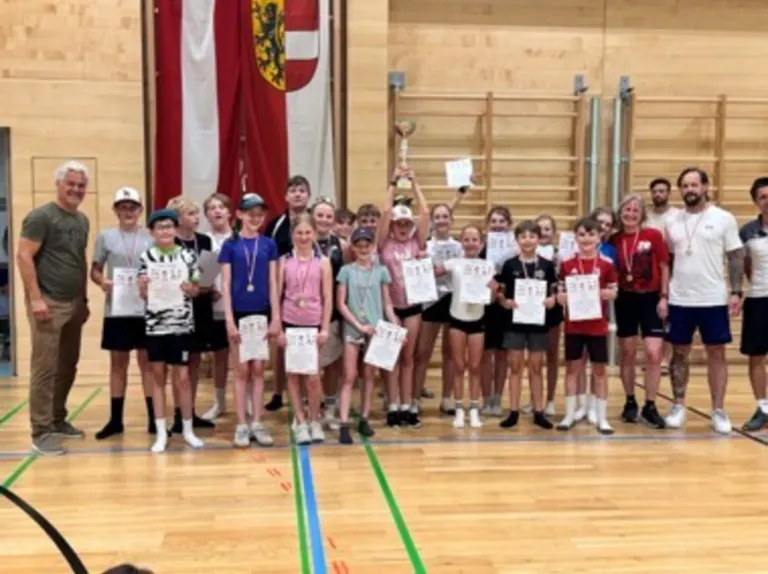 Eine Gruppe von Kindern und Jugendlichen steht auf einem Basketballplatz, hält Urkunden, trägt Medaillen und hält einen Pokal. Hinter ihnen wird eine Flagge gezeigt.
