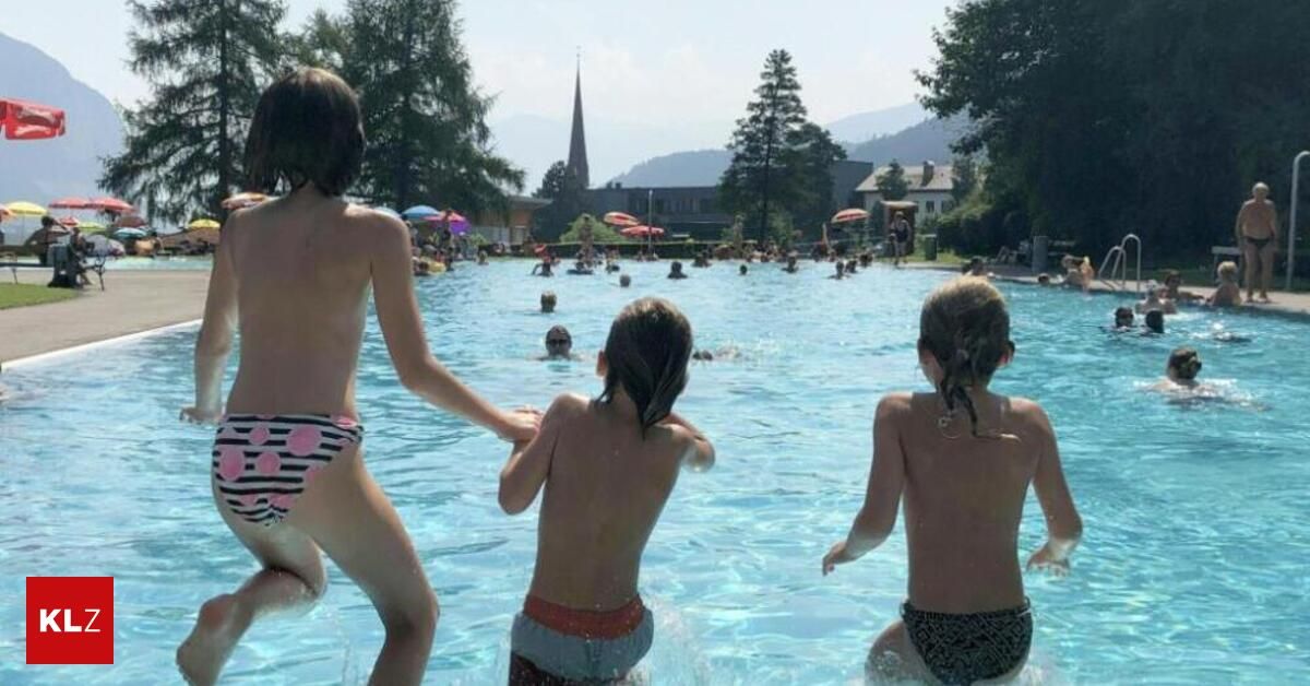 Mehrere Kinder spielen in einem großen Freibad, umgeben von Bäumen und Gebäuden. Es gibt Schwimmende und andere, die am Rand stehen.