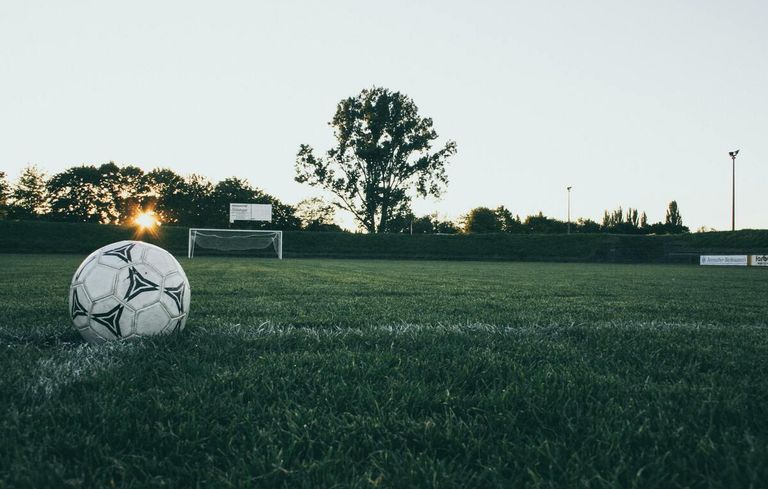 Ein Fußball liegt auf einem Feld mit einem Tornetz und einem Schild im Hintergrund. Das Feld ist von Bäumen umgeben und der Himmel ist klar.