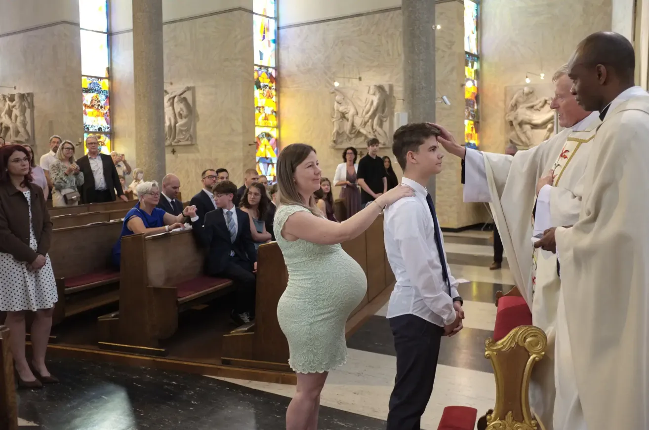 Eine schwangere Frau steht neben einem jungen Mann in einer Kirche. Der Priester segnet den jungen Mann. Die Gemeinde sitzt auf Kirchenbänken.