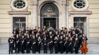Ein Orchester von Musikern in Uniform steht auf den Stufen eines großen Gebäudes und hält verschiedene Musikinstrumente in den Händen. Das Gebäude hat kunstvolle Fenster und ein Schild über dem Eingang, das 'Gluck auf' lautet.