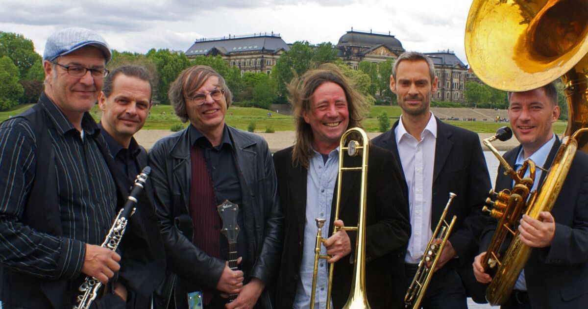 Eine Gruppe von Männern lächelt und posiert für ein Foto, während sie Musikinstrumente halten. Hinter ihnen steht ein großes Gebäude und Bäume.