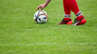Ein Fußballspieler berührt einen Fußball auf einem grünen Feld und trägt Adidas-Schuhe.