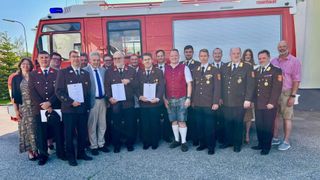 Eine Gruppe von Menschen in Uniform steht vor einem roten Feuerwehrauto, hält Urkunden und posiert für ein Foto.