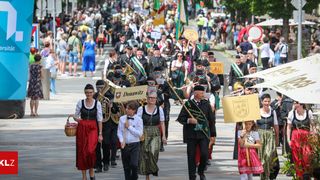Ein Straßenumzug mit einer Blaskapelle, Musikern, die Blechblasinstrumente spielen, und Zuschauern. Einige Teilnehmer tragen traditionelle Kleidung und tragen Banner.