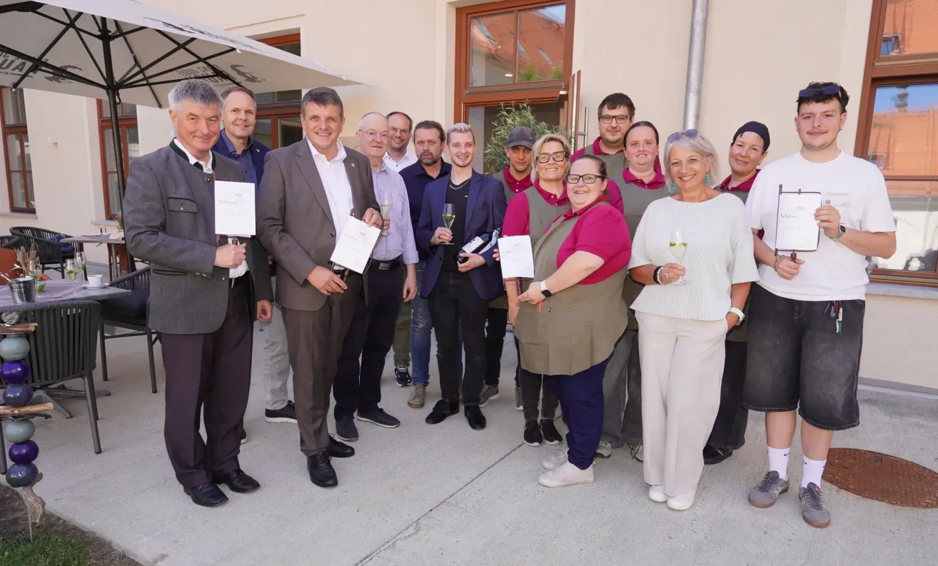 Eine Gruppe von Menschen steht draußen und hält Weingläser und Urkunden in der Hand. Sie sind geschäftlich gekleidet und scheinen ein Ereignis zu feiern. Hinter ihnen steht ein Gebäude mit Glasfenstern.