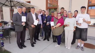 Eine Gruppe von Menschen steht draußen und hält Weingläser und Urkunden in der Hand. Sie sind geschäftlich gekleidet und scheinen ein Ereignis zu feiern. Hinter ihnen steht ein Gebäude mit Glasfenstern.