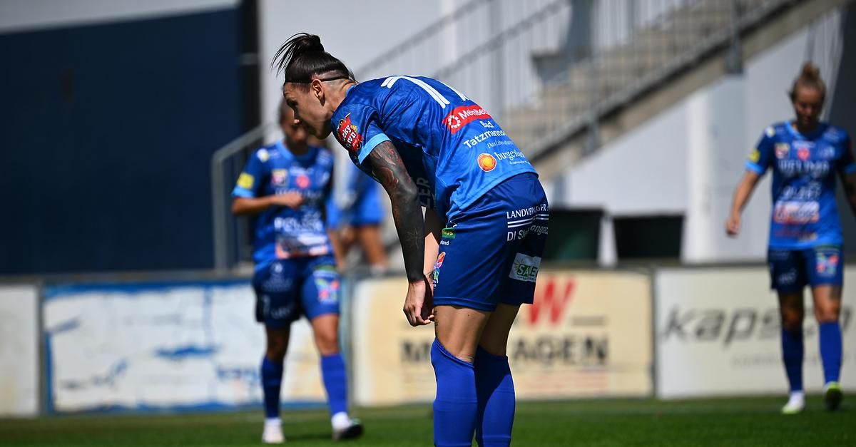 Ein Fußballspieler in einem blauen Trikot steht auf einem Feld, schaut nach unten und hat die Hände auf den Knien. Im Hintergrund geht ein anderer Spieler.