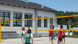 Eine Gruppe von Menschen spielt Volleyball auf einem Sandplatz. Das Netz ist gelb, und sie tragen Teamuniformen. Ein Mann ist in der Luft und bereit, den Ball zu schlagen.