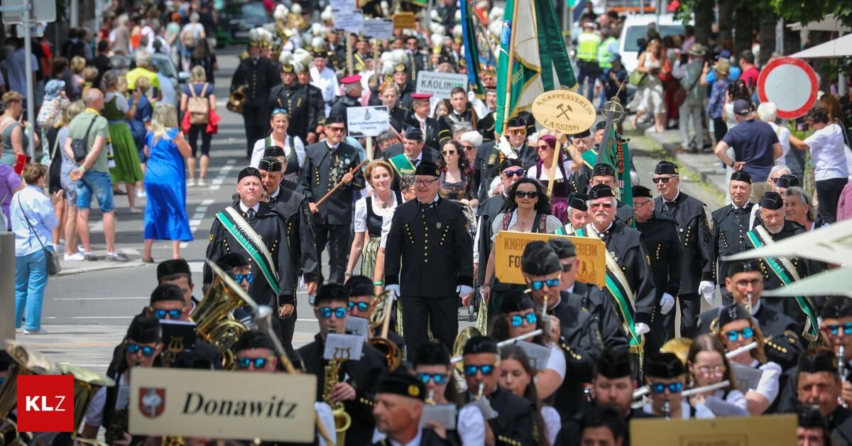 Ein Umzug in einer Stadt mit Blaskapellen und Menschen in traditionellen Kostümen. Ein Schild sagt 'Donawitz' und die Teilnehmer tragen Flaggen. Die Veranstaltung scheint eine festliche Feier zu sein.