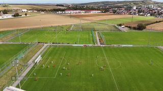 Ein großes Fußballfeld mit mehreren Spielern in roten und weißen Uniformen, die ein Spiel spielen. Das Feld ist von Gras umgeben und hat mehrere Tore. In der Ferne gibt es Gebäude und ein großes Feld.