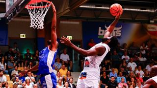 Zwei Basketballspieler in einem Spiel. Einer springt zum Dunken, der andere verteidigt mit erhobenen Händen. Das Spielfeld hat ein Publikum, einen Korb und ein blaues Banner.