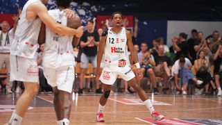 Basketballspieler in weißem Trikot mit Nummer 12 auf dem Spielfeld, schreit, mit einem anderen Spieler und jubelnden Zuschauern im Hintergrund.