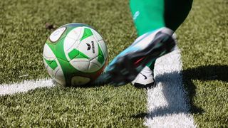 Ein Fußball mit der Aufschrift Erima liegt auf dem Boden. Ein Fuß in einem Nike-Schuh ist neben dem Ball positioniert.