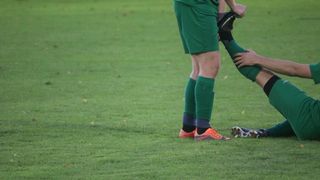 Ein Fußballspieler in grüner Uniform hilft einem anderen Spieler mit einer Fußverletzung auf einem grünen Rasen.
